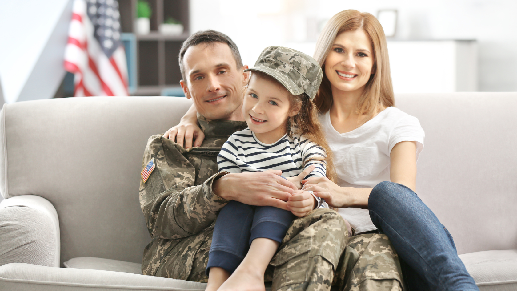 A smiling military family sitting together on a couch, with a service member in uniform holding his young daughter in his lap while the mother sits beside them. An American flag is visible in the background, creating a warm and patriotic scene.