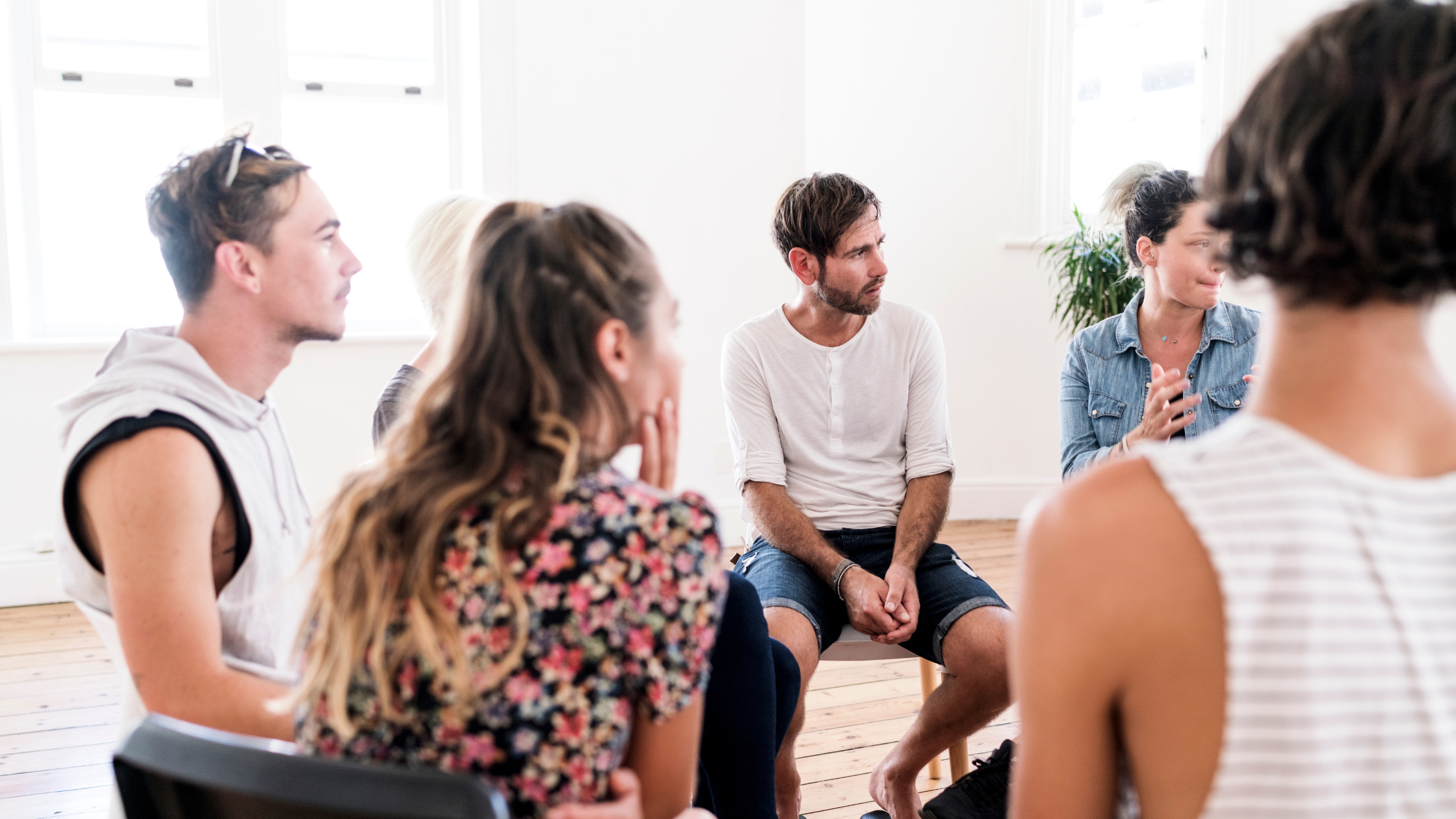 A small group of adults sits in a bright, airy room during a therapy or support group session. They are arranged in a circle, listening attentively as one woman speaks, creating a calm and supportive atmosphere.