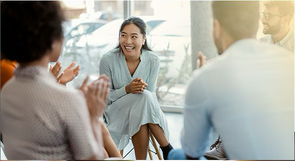 A woman smiling and engaging in a supportive group therapy session while others sit in a circle and clap around her.