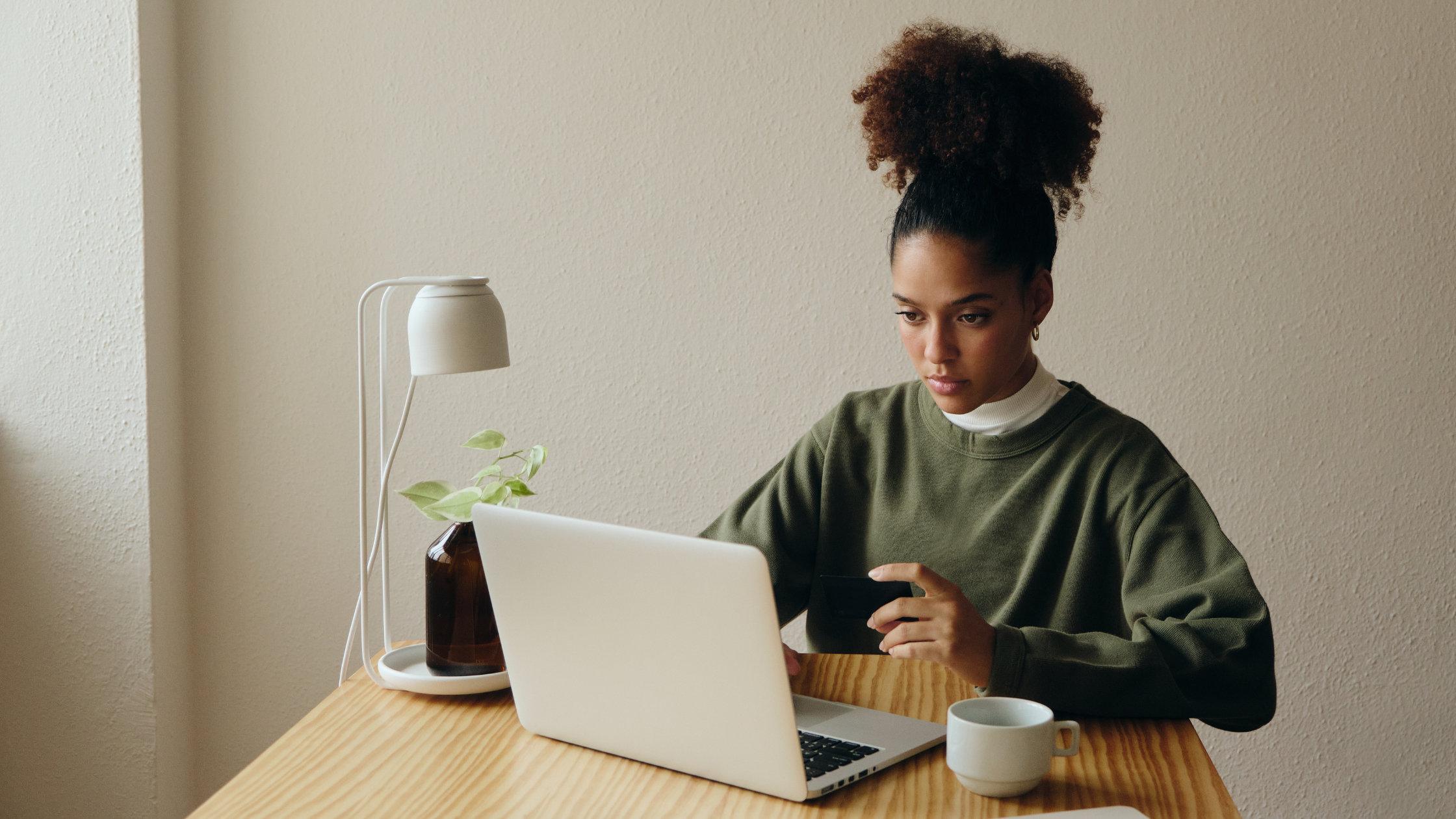 Woman sitting at a desk with a laptop, holding a credit card and looking focused, symbolizing the rise of digital tools like AI in therapy. A plant, coffee mug, and lamp add a calm, modern workspace feel.