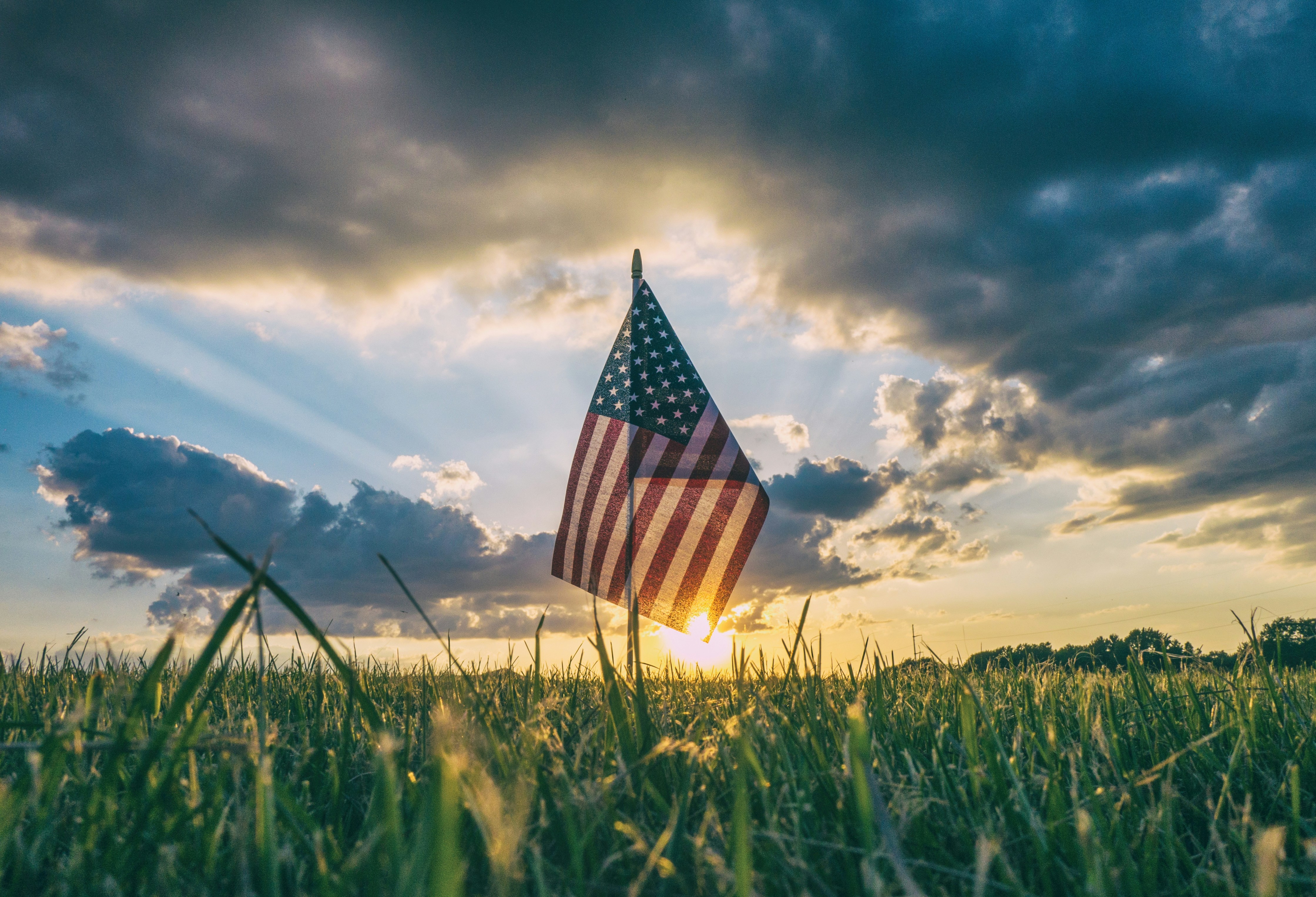 "American flag in a grassy field at sunset with dramatic clouds and sun rays, symbolizing Independence Day and reflection on the meaning of freedom."