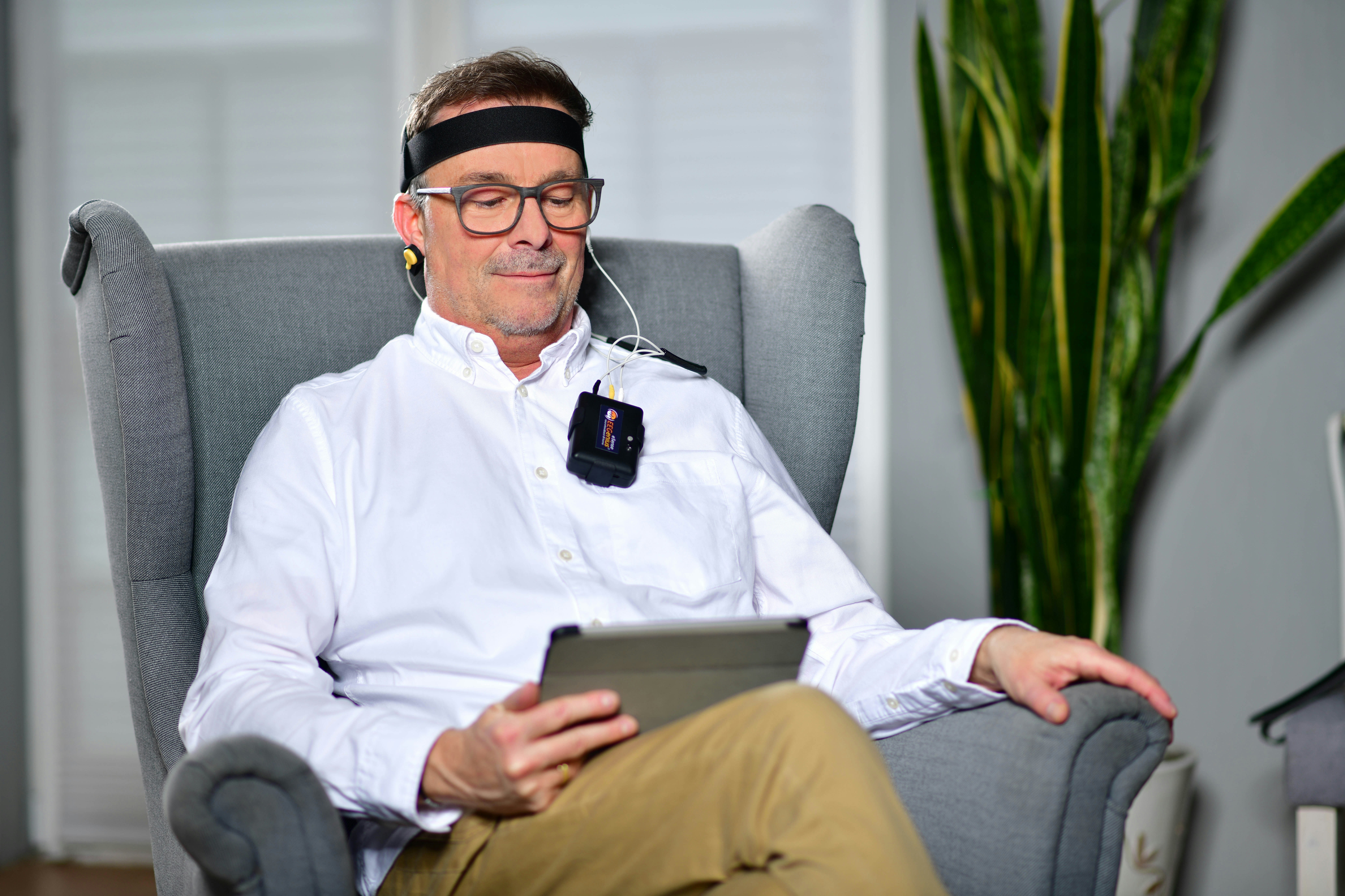 Man sitting in a comfortable chair using a neurofeedback device with electrodes attached to his head and earphones, looking at a tablet, appearing calm and focused.